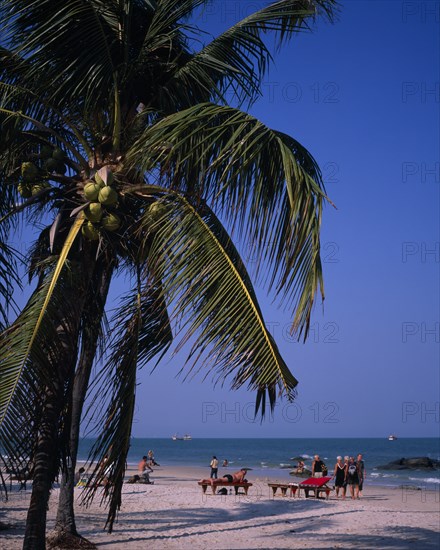 THAILAND, Hua Hin, Western tourists on Hua Hin beach with distant boats on the water and coconut palms in foreground casting shadows across the sand.