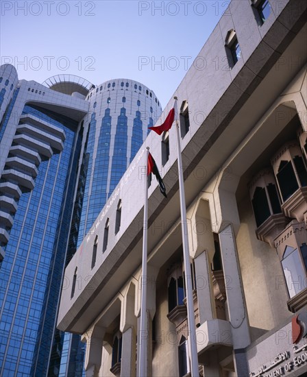 UAE, Dubai, "Two flag poles in front of contrasting styles of architecture, modern, mainly glass skyscraper behind facade of building in more traditional style."