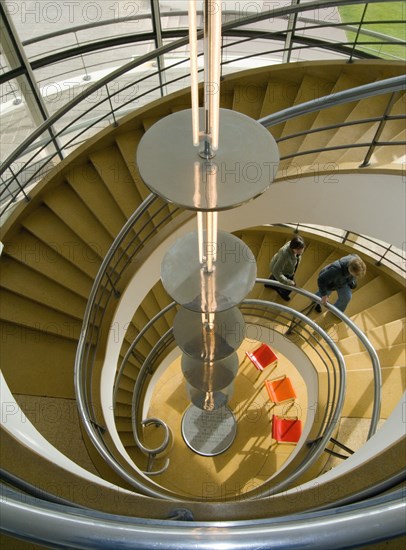 ENGLAND, East Sussex, Bexhill-on-Sea, The De La Warr Pavilion. Interior view down the helix  staircase with chrome Bauhaus globe lamps. Visitors walking the stairs. Three colourful chairs seen at the bottom.