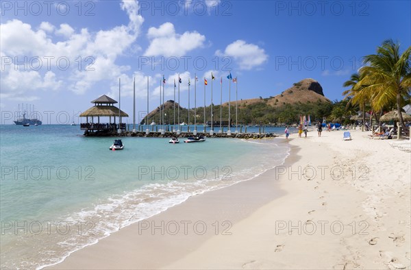 WEST INDIES, St Lucia, Gros Islet , Tourists on the beach at Sandals Grande St Lucian Spa and Beach Resort hotel beside a wooden jetty with Pigeon Island National Historic Park beyond