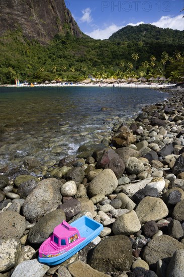 WEST INDIES, St Lucia, Soufriere , Val des Pitons The white sand beach at the Jalousie Plantation Resort Hotel with the volcanic plug of Petit Piton beyond and tourists on sunbeds beneath palapa sun shades with a childs plastic boat marooned on rocks in the foreground