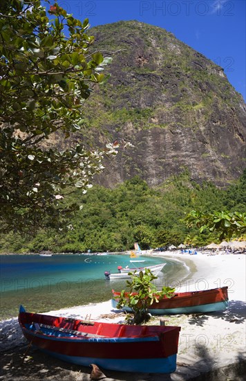 WEST INDIES, St Lucia, Soufriere , Val des Pitons The white sand beach at the Jalousie Plantation Resort Hotel with the volcanic plug of Petit Piton beyond and tourists on sunbeds beneath palapa sun shades with colourful fishing boats under a tree in the foreground