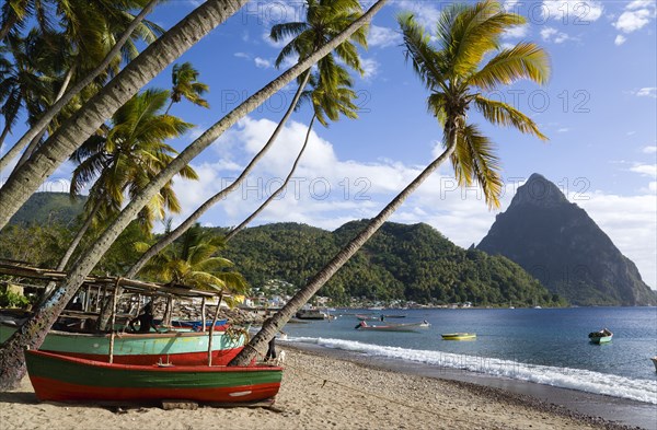 WEST INDIES, St Lucia, Soufriere, Fishing boats on the beach lined with coconut palm trees with the town and the volcanic plug mountain of Petit Piton beyond