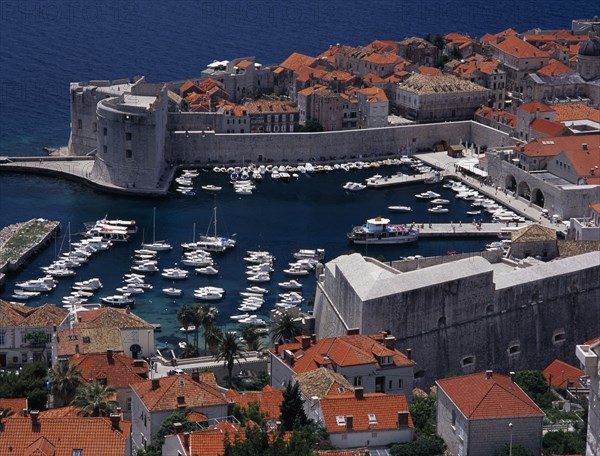 CROATIA, Dalmatia, Dubrovnik, Elevated view over the Old City Harbour with fortified walls. Yachts moored in marina surrounded by buildings with terracotta tiled rooftops