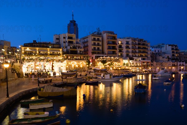 MALTA, Saint Julians, Saint Julians Bay waterfront at dusk with illuminated restaurants in front of apartments and the Hilton Tower office complex with fishing boats in the harbour in the foreground