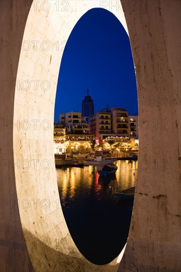 MALTA, Saint Julians, Saint Julians Bay waterfront at dusk with illuminated restaurants in front of apartments and the Hilton Tower office complex with fishing boats in the harbour in the foreground seen through a spherical opening