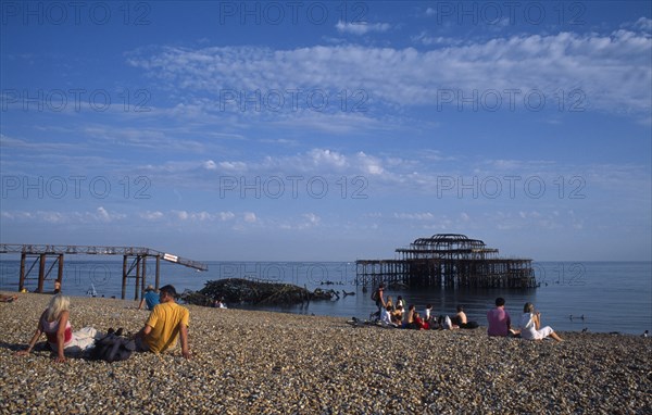 ENGLAND, East Sussex, Brighton, Groups of people sitting on the beach by the ruined West Pier in late afternoon sunshine.