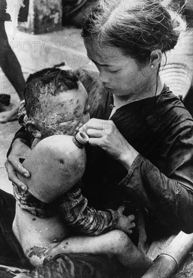 VIETNAM, South, Cam Che, Badly burned Vietnamese baby caught in bursting napalm bomb between US Marines and North Vietnamese taking water from a soldier’s water bottle on his mother’s knee.