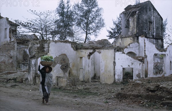 VIETNAM, North, War, Woman carrying basket of vegetables on her head passing ruins of war damaged building.