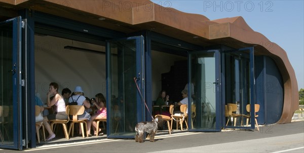 ENGLAND, West Sussex, Littlehampton, Customers sitting at tables inside the East Beach Cafe designed by Thomas Heatherwick. Dog tied to glass door waiting outside