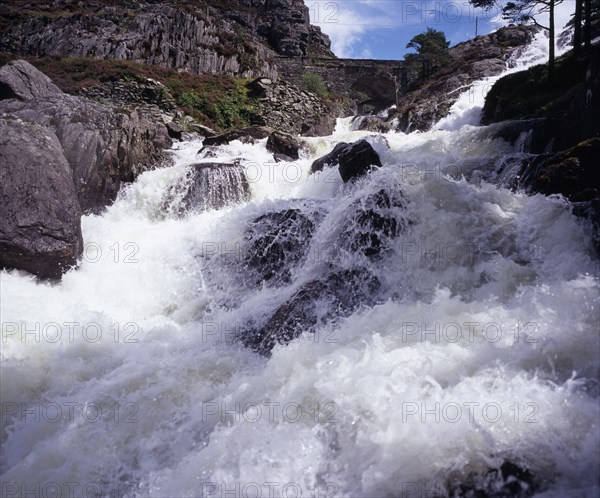 WALES, Gwynedd, Snowdonia National Park, Ogwen Falls and Nant Ffrancon Pass.   White foaming water tumbling over rocks with arched stone bridge in background.