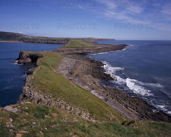 WALES, Swansea, Gower Peninsula, View east from the Worm’s Head towards the mainland.  Sloping ridge of cliff extending from mainland with waves breaking against side.