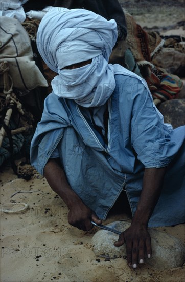ALGERIA, Sahara Desert, Tuareg, "Tuareg man sharpening tool on rock, wearing blue taguelmoust covering face and head except for eyes. In Tuareg societies men are veiled rather than women.   "