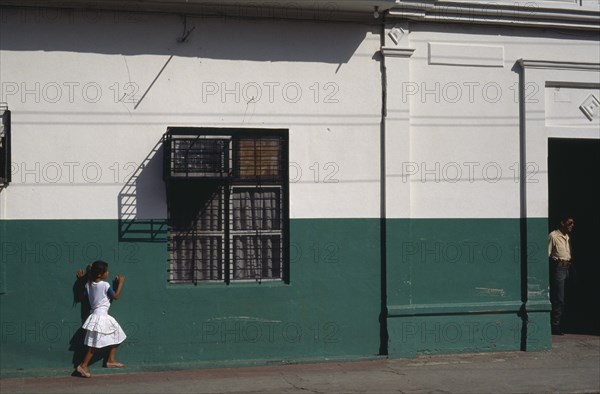 NICARAGUA, Leon, Street scene with little girl in white outside building painted dark green and white.  Metal window grill casting shadow above and man standing in doorway on right.