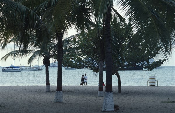 VENEZUELA, Estado Anzoategui, Puerto La Cruz, Couple on beach looking out over water and moored yachts framed by palms and other trees in foreground.