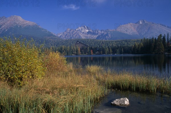 SLOVAKIA, Carpathian Mtns, High Tatras Mtns, Strbske Pleso.  View across lake towards distant chalet and mountains beyond with surrounding trees and reeds and vegetation in foreground in Autumn colours.