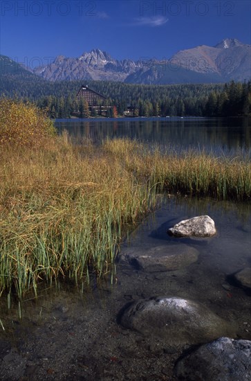 SLOVAKIA, Carpathian Mtns, High Tatras Mtns, Strbske Pleso.  View across lake towards distant chalet and mountains beyond with surrounding trees and reeds and vegetation in foreground in Autumn colours.