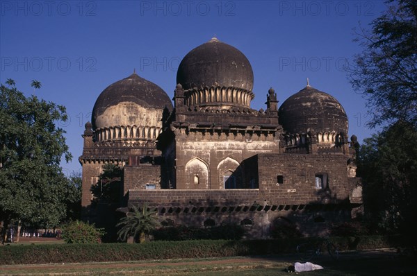 INDIA, Karnataka, Bijapur, Unidentified old building in Islamic architectural style with three domes set in gardens or green space with person asleep beside bicycle in foreground.