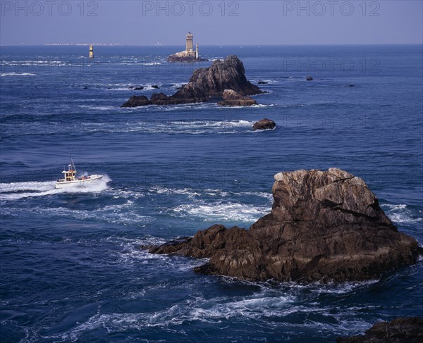FRANCE, Brittany, Finistere, "Pointe du Raz.  Jagged outcrops of offshore rock protruding from sea with small fishing boat facing oncoming tide, light house and Ile de Sein skyline."
