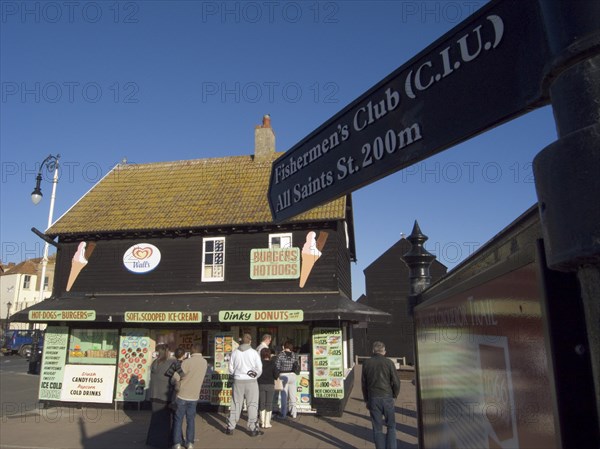 ENGLAND, East Sussex, Hastings, Customers queuing at a seaside take away. A sign with directions to the Fisherman’s club in the foreground