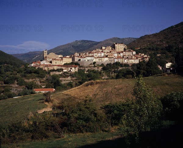 FRANCE, Languedoc-Roussillon, Pyrenees-Orientales, Mosset. View towards hillside village from the east with narrow houses with painted window shutters and red tiled rooftops. Backdrop of rocky hills scatteres with trees.