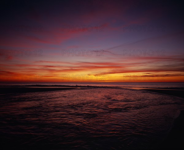 FRANCE, Aquitaine, Landes, Cap de L’Homy.  Red and orange sunset sky over Atlantic coastline with windswept clouds and rippled water surface.