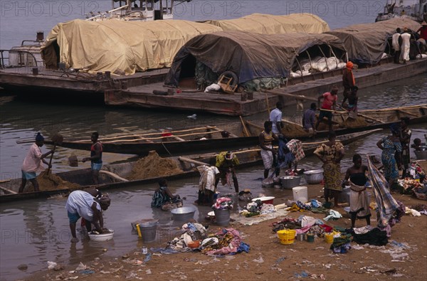 SENEGAL, Niger River, Women washing clothes in river from bank with barges and wooden canoes transporting soil and other goods behind.
