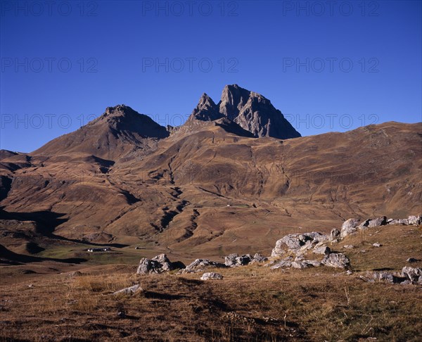 FRANCE, Aquitaine, Pyrenees Atlantiques, Pic du Midi d’Ossau (2884 m) in Autumn. Eroded mountain-side and scattered houses with narrow winding road across centre-ground. Rocks in foreground.