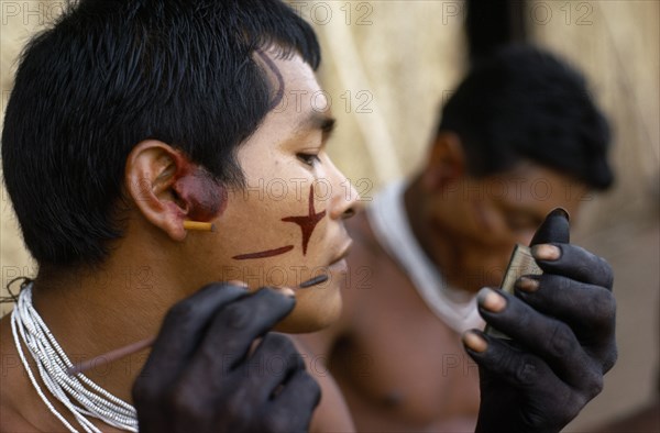 COLOMBIA, North West Amazon, Tukano Indigenous Tribe, "Barasana men (sub group of Tukano) decorating their faces with red Achiote facial paint for ceremonial dance/festival Hands, wrists blackened with dark purple/black juice from We leaves Wearing necklaces of small white glass trade beads "
