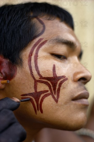 COLOMBIA, North West Amazon, Tukano Indigenous Tribe, Barasana man (sub group of Tukano) decorating his face with red Achiote facial paint for manioc festival and dance ceremonial. Hands wrists and body decoration with dark purple/black juice from We leaves