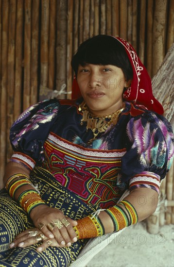 PANAMA, San Blas Islands, Kuna Indigenous Tribe, "Portrait of a Kuna Indian woman in hammock  with gold jewelry including gold nose ring and rings on fingers, bead design arm bands, wearing brightly coloured layered applique traditional Mola with fine bird design  beads on arms and a gold nose ring. Cuna"