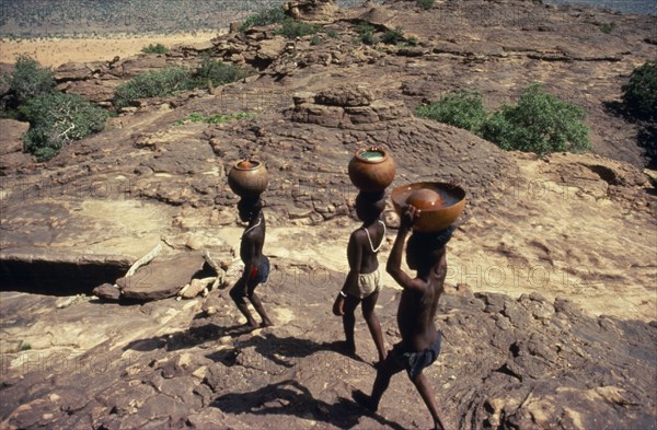 20088820 MALI Pays Dogon People Dogon boys carrying water pots on their heads over rocky terrain.