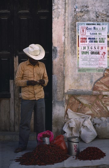 MEXICO, Oaxaca, Man standing beside crumbling plaster wall with pile of red chillies for sale at his feet.
