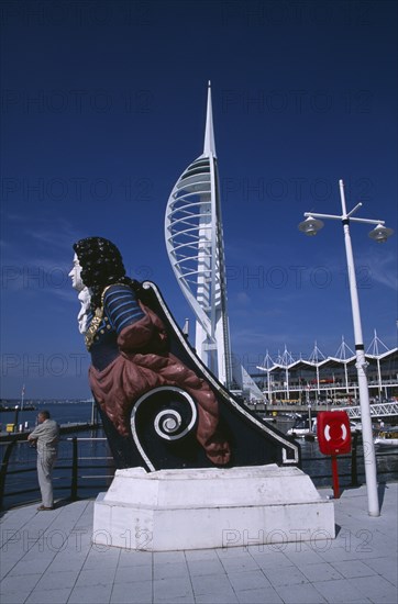 ENGLAND, Hampshire, Portsmouth, Gunwharf Keys. The Spinnaker Tower with figurehead in the foreground and a man standing on waterfront promenade looking out across the harbour