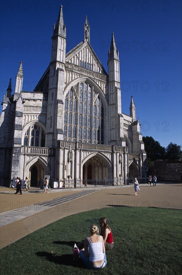 ENGLAND, Hampshire, Winchester, Winchester Cathedral exterior seen from the gardens with two girls sat on the grass in the foreground.