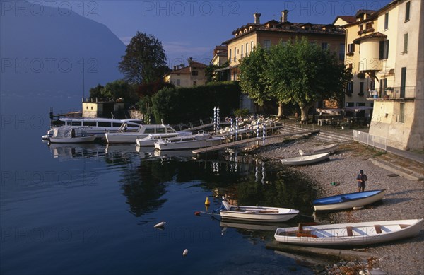 ITALY, Lombardy, Lake Como, Sala Comacina.  Fishing village with motor boats moored against wooden jettys extending from pebble beach overlooked by pastel coloured facades of houses and cafe
