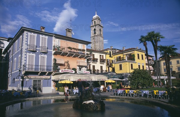 ITALY, Piedmont, Lake Maggiore, Pallanza.  Bar and cafe with people sitting at outside tables in  square with central circular fountain.  Pastel coloured building facades and bell tower part seen above