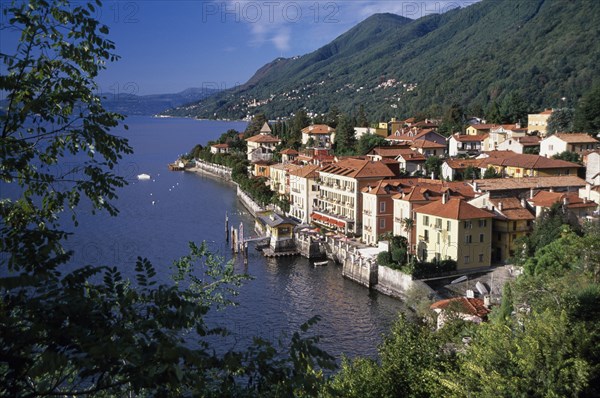 ITALY, Piedmont, Lake Maggiore, Cannero Riviera.  Waterside buildings with red tiled rooftops at foot of densely wooded hillside with view across lake part framed by tree branches.