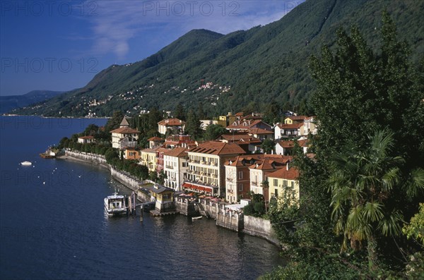 ITALY, Piedmont, Lake Maggiore, Cannero Riviera.  Waterside buildings with red tiled rooftops at foot of densely wooded hillside with ferry moored against short jetty and view across lake.