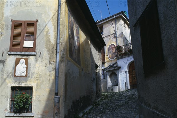 ITALY, Lombardy, Arcumeggia, Narrow cobbled street with fresco decoration and plaster relief of the Virgin and Child on the exterior walls of the buildings and drinking fountain at top.