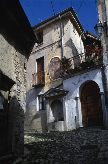 ITALY, Lombardy, Arcumeggia, Narrow cobbled street with fresco decoration on exterior wall of building and drinking fountain below beside arched doorway with balcony and flowers above.