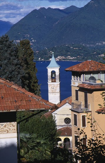 ITALY, Piedmont, Lake Maggiore , Stresa.  Overhanging red tiled rooftops of town buildings with church bell and clock tower in centre.  View across lake towards mountain behind.