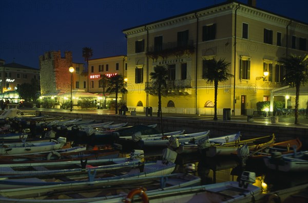 ITALY, Veneto, Lake Garda, Bardolino.  Harbour and moored boats at night with waterside bars and hotels and illuminated street lights and signs.