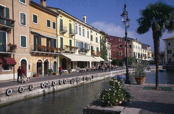 ITALY, Veneto, Lake Garda, Bardolino.  Harbour and line of waterside bars and cafes painted in pastel colours and with awning pulled out over outside tables.  Moored boats in foreground.