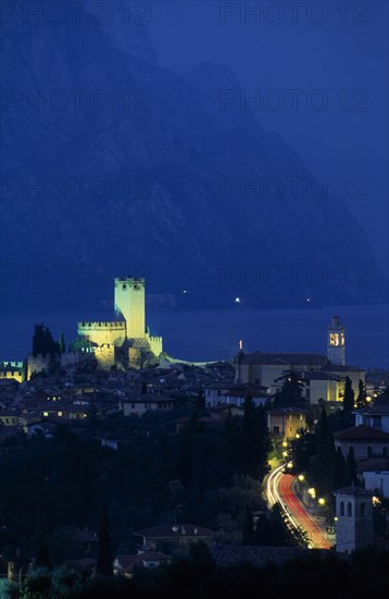 ITALY, Veneto, Lake Garda , Malcesine.  Tiled rooftops of town and castle illuminated at night with lake beyond.  Coloured light trails from cars on road in foreground.