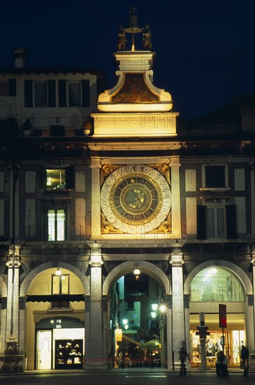 ITALY, Lombardy, Brescia, "Astronomical clock in Piazza della Loggia illuminated at night with brightly lit shop windows framed by archways of colonnade below.  People, street lamps and light trail."
