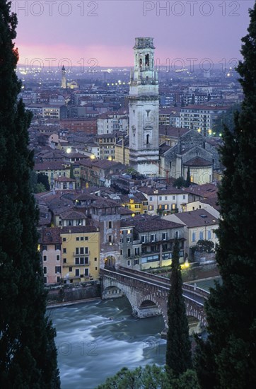 ITALY, Veneto, Verona, "Cityscape at sunset showing tiled rooftops, Duomo bell tower and bridge over the Adige River part framed by tall coniferous trees.  Pale pink/purple sky and city lights."