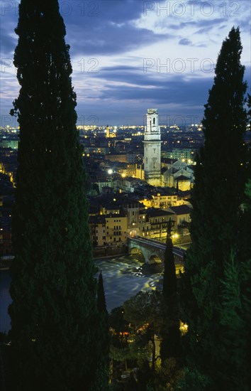 ITALY, Veneto, Verona, "Cityscape illuminated at night showing tiled rooftops, Duomo bell tower and bridge over the Adige River part framed by tall coniferous trees.  Windswept evening sky."