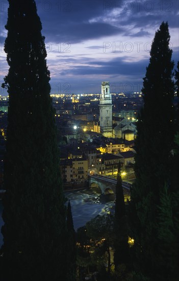 ITALY, Veneto, Verona, "Cityscape illuminated at night showing tiled rooftops, Duomo bell tower and bridge over the Adige River part framed by tall coniferous trees.  Windswept evening sky."