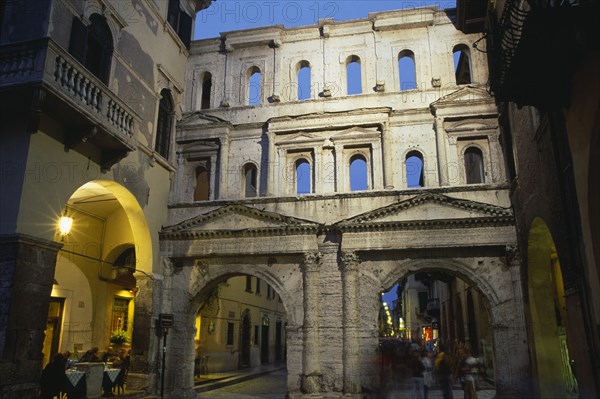 ITALY, Veneto, Verona, Street scene and Roman gateway at night with group of people framed in archway.  Light from street lamps and shop windows and people at restaurant tables on left.